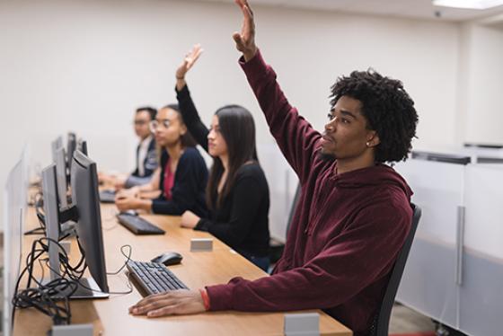 Four students sit at computer desks in class, two raise their hands.