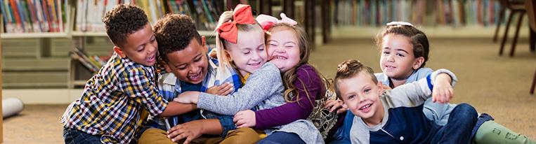 Six young children sit in a line on the floor of a library and hug each other.