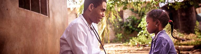 A health care provider wearing a stethoscope sits facing a young girl and holds her hand outside her home.