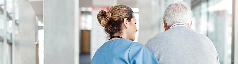 A health care provider holds an older man's arm to help him walk down a hallway.