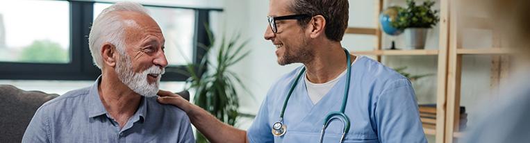 A health care provider sitting next to a patient places his hand on the patient's shoulder and smiles.