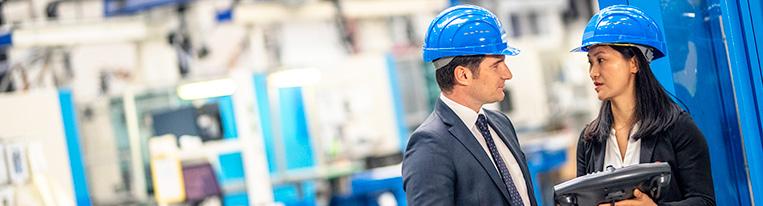 A man and woman wearing business suits and hard hats have a discussion inside a warehouse.