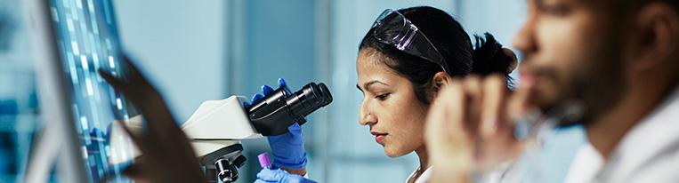 A woman in a white lab coat uses a microscope while a man in a lab coat uses a computer touchscreen.