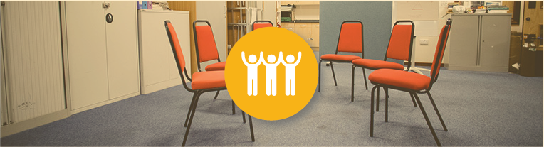 An icon of 3 people overlays a circle of red chairs in a community center.