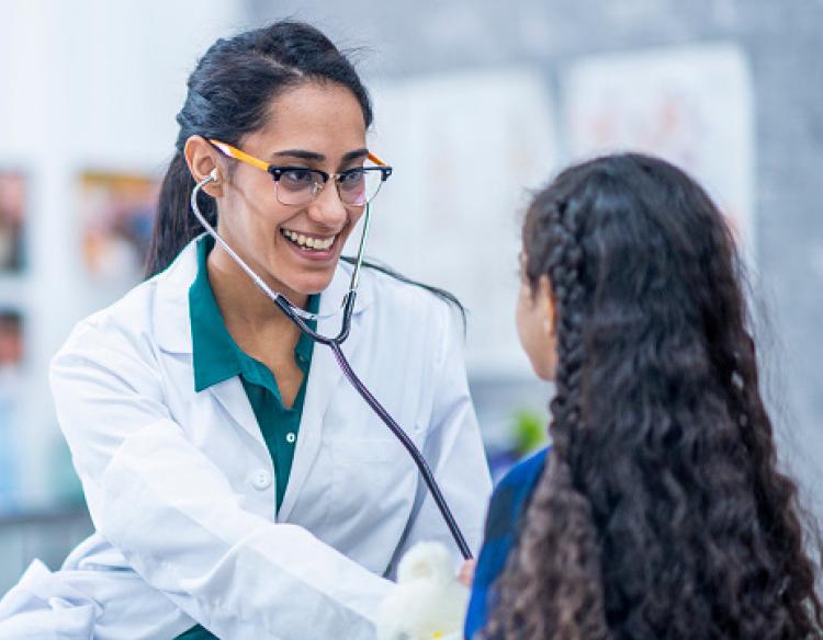 A doctor with long dark hair in a ponytail, glasses, and a white lab coat smiles and checks a child’s  vital signs using a stethoscope.