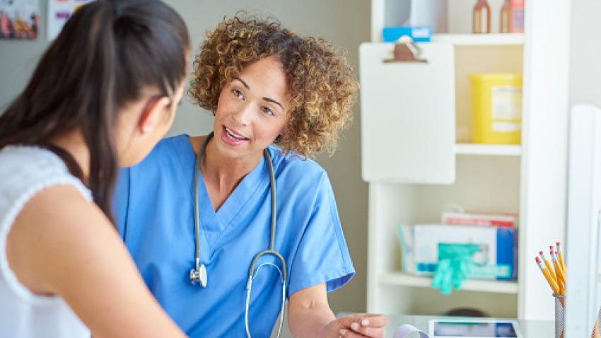 A nurse with dark curly hair wearing blue scrubs talks to a patient.