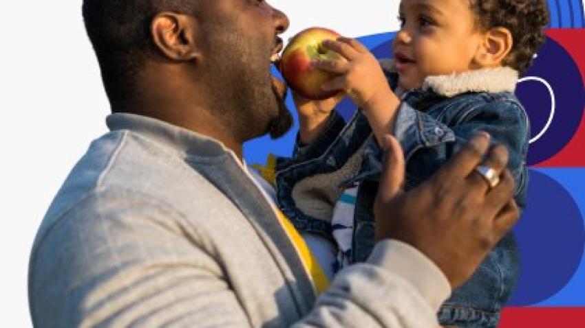 A dad holds his young child while the child holds an apple up to his mouth for him to bite into.