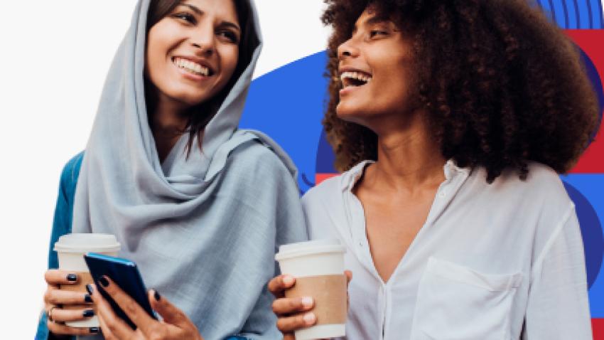 Two women walking together and smiling while holding coffee. One woman wears a headscarf while the other woman has brown tight curly hair.